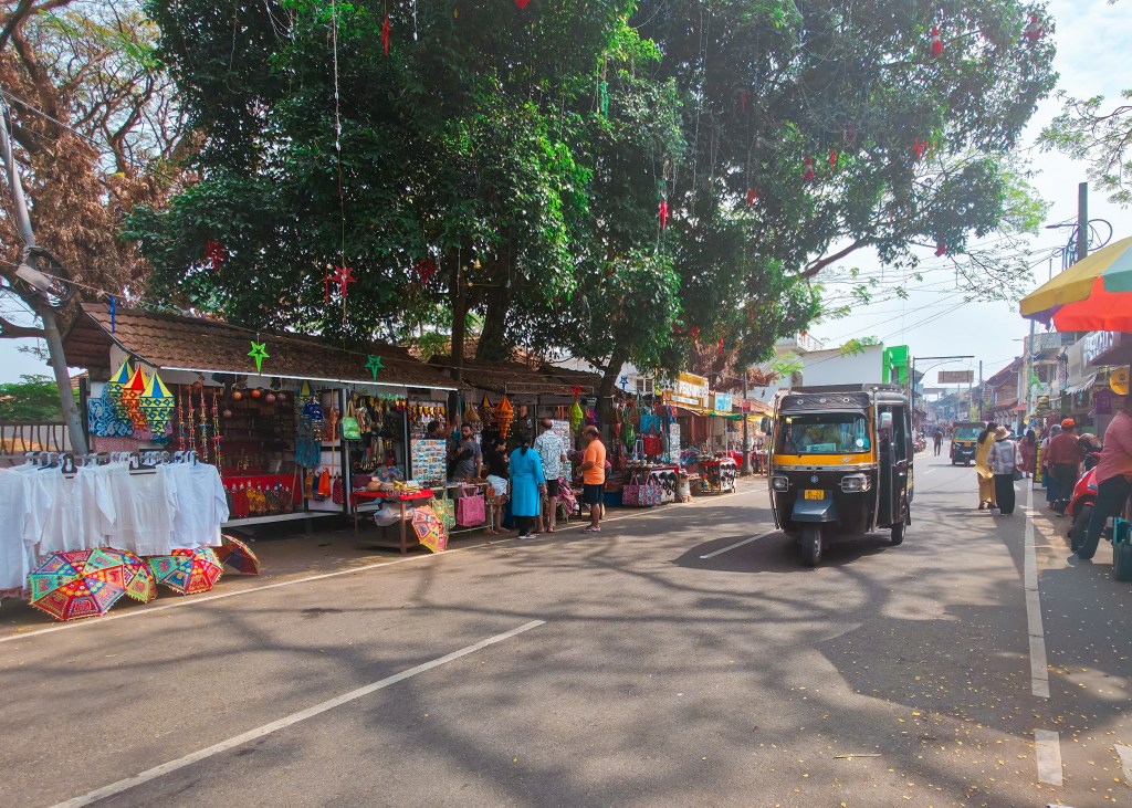Street market lined with small stalls selling colourful lanterns, umbrellas, and souvenirs beneath large trees, while an auto rickshaw drives along the road and people browse the stalls.