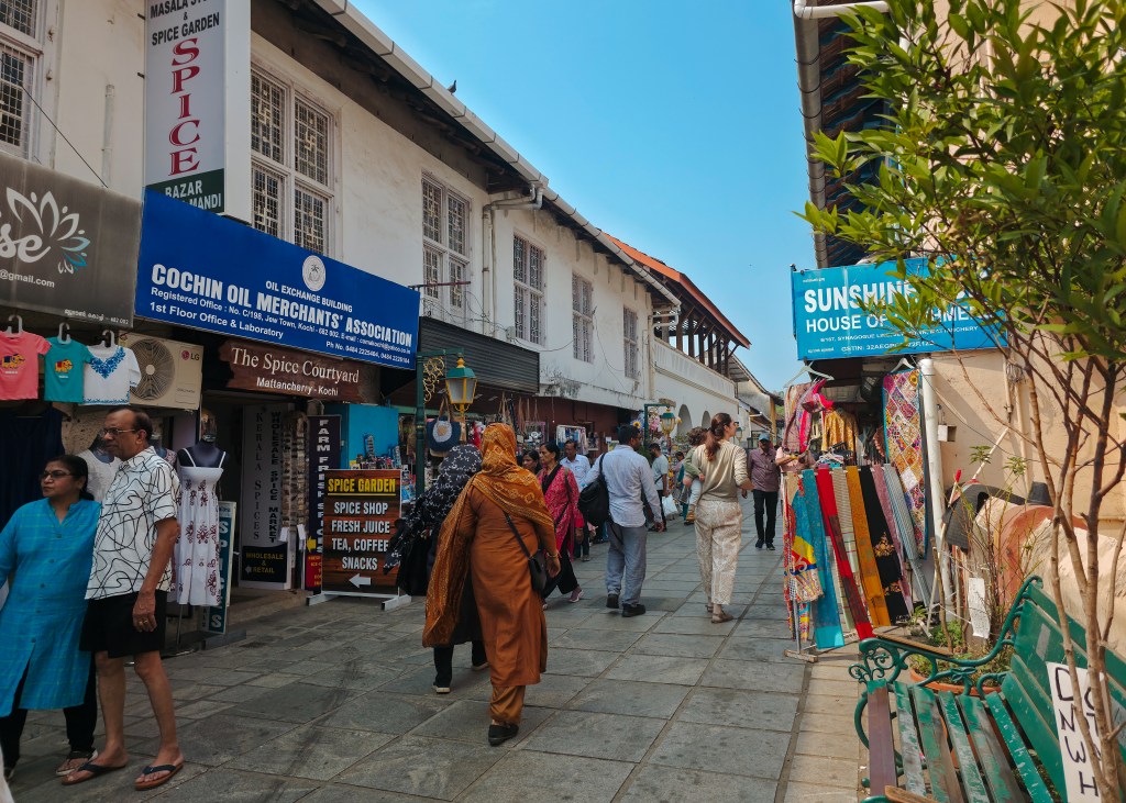 Pedestrian street in Jew Town, Kochi with small shops selling spices, clothing and souvenirs as people walk between storefronts under a clear blue sky.