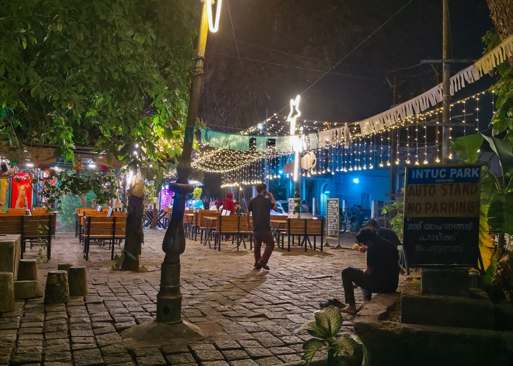 Outdoor restaurant courtyard at night lit with string lights and lanterns, with wooden tables and chairs set on a cobblestone surface as people sit and walk through the space.