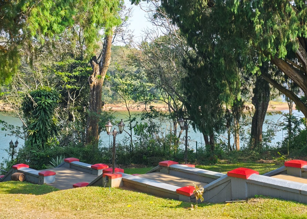 Garden steps with red-painted edges lead down toward a lakeside, with trees and lampposts along the water’s edge.