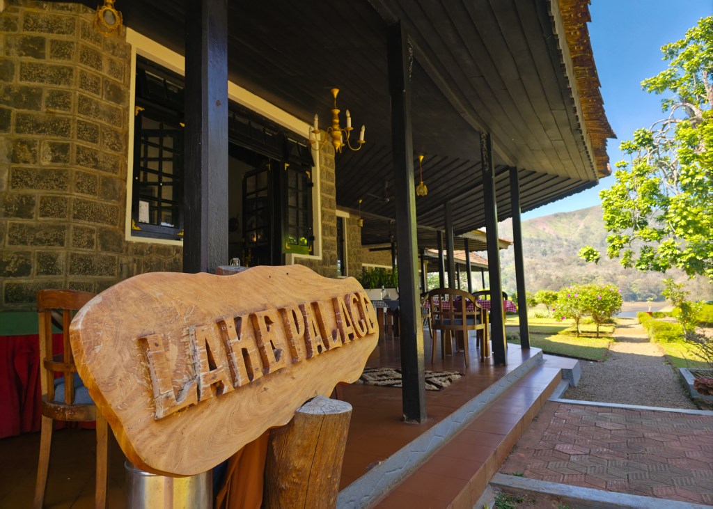 Wooden sign reading “Lake Palace” sits on a veranda of a stone building with black pillars, overlooking a garden and water with hills in the background.
