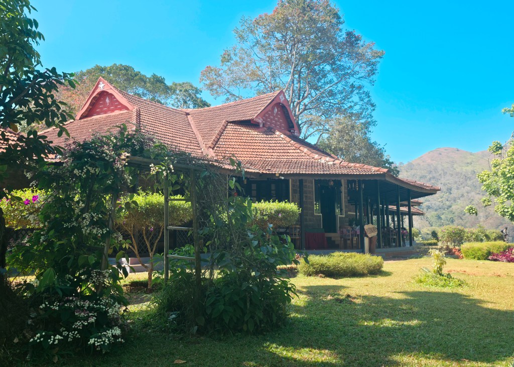 Traditional building with a tiled roof set among gardens, with hills in the background under a clear sky.