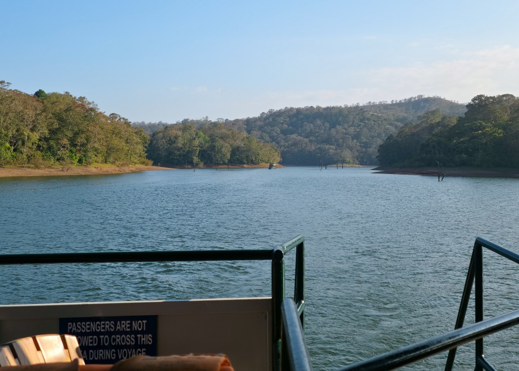 View from a boat across a wide lake toward forested hills under a clear sky, with a railing in the foreground.